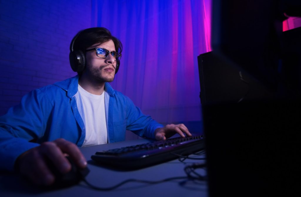 Young man wearing blue light glasses while playing video games on computer at night