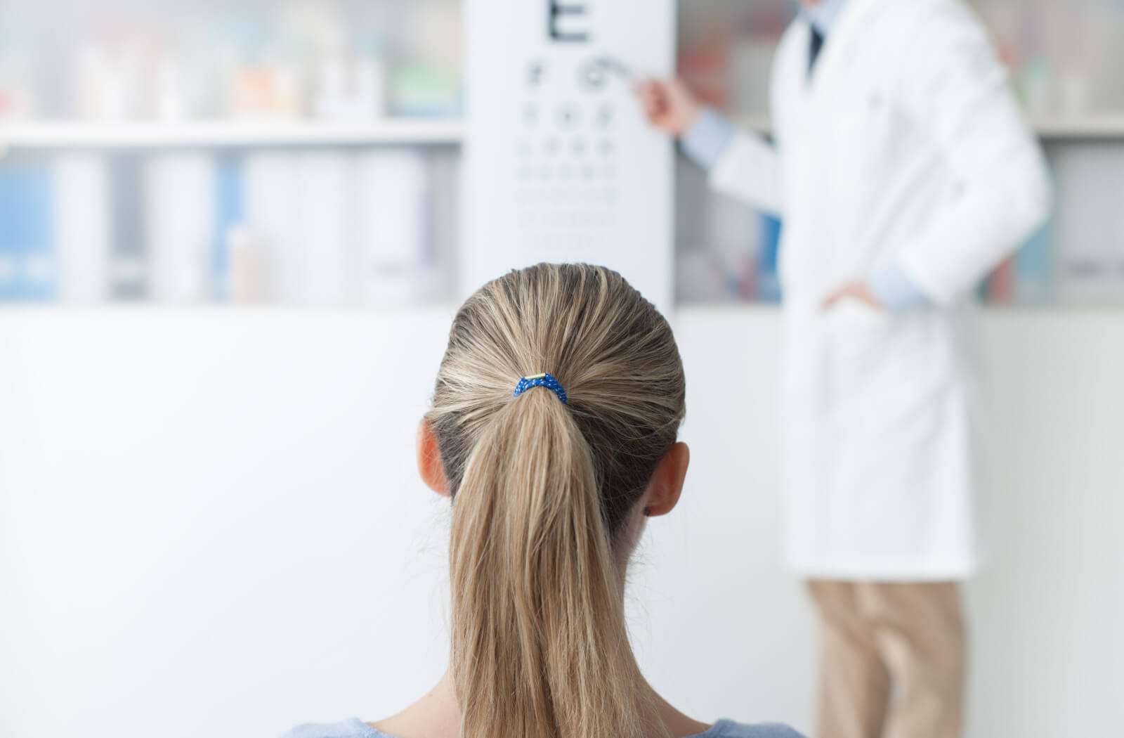 A young woman in an optical clinic undergoing an acuity test, an optician is pointing a letter from the chart while the patient is calling out the letter from a far,