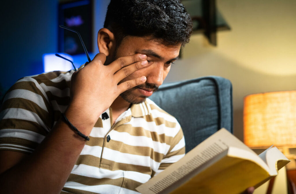 A young man reading a book while rubbing his right eye with his right hand and holding his glasses with the same hand.