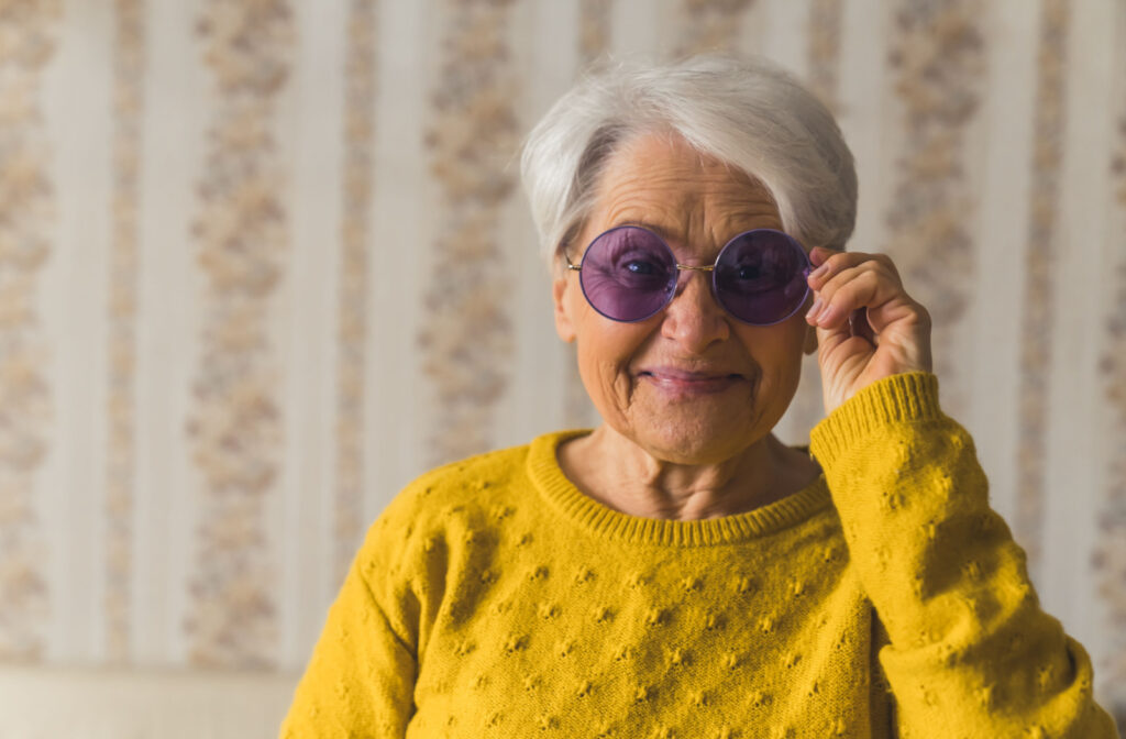 A senior woman in a yellow sweater wearing sunglasses indoors to help recover from a cataract surgery.