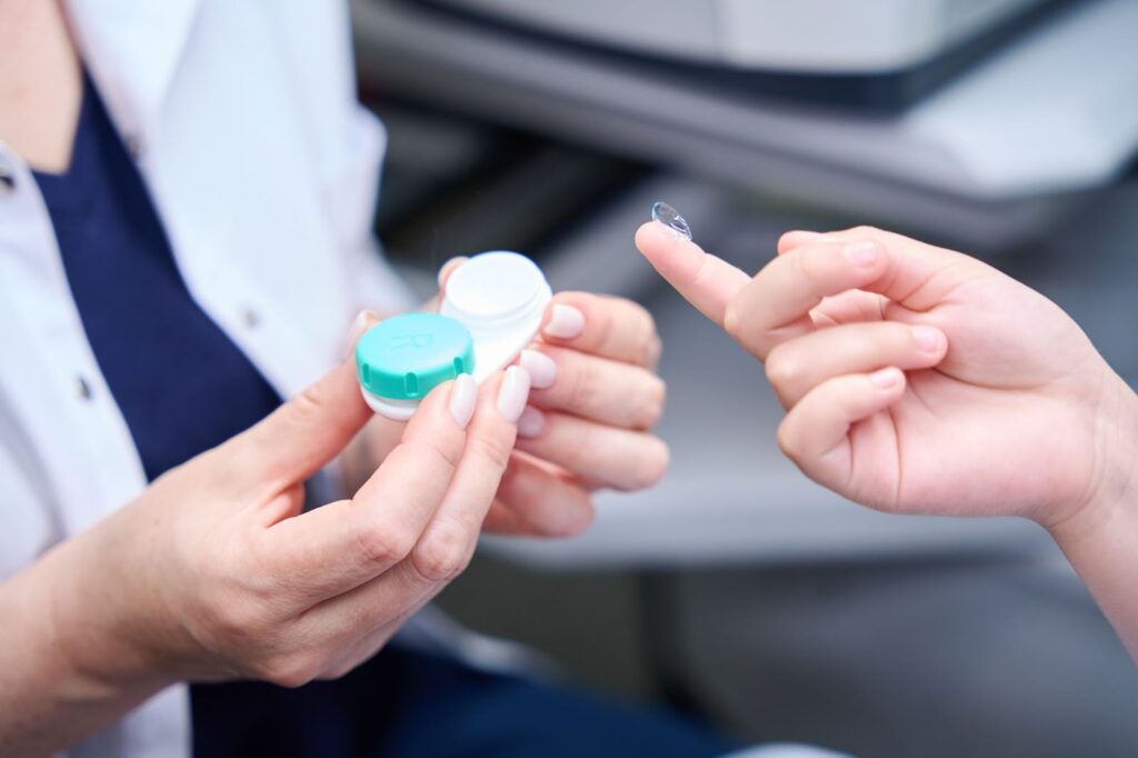 A close up of an eye doctor's hands holding an open contact lens case and a patient's finger with a contact lens.